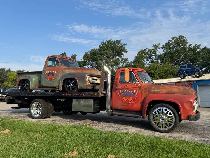 Vintage Ford F-600 flatbed carrying a patina Ford F-100, both wearing Midwest Custom Trucks branding and custom wheels outside the Moberly shop
